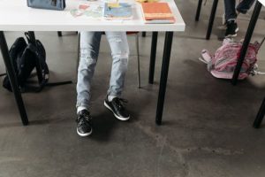 A student wearing sneakers and ripped jeans sits under a desk in a classroom.